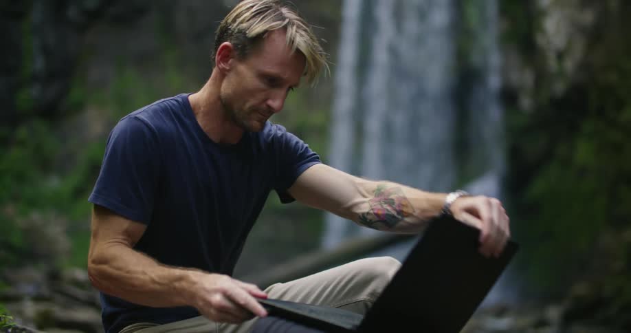 Man sitting on riverbank near waterfall opens laptop and starts typing