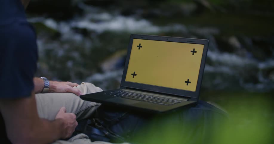 A man is sitting on a ledge near a stream looking at a laptop computer with a blank screen