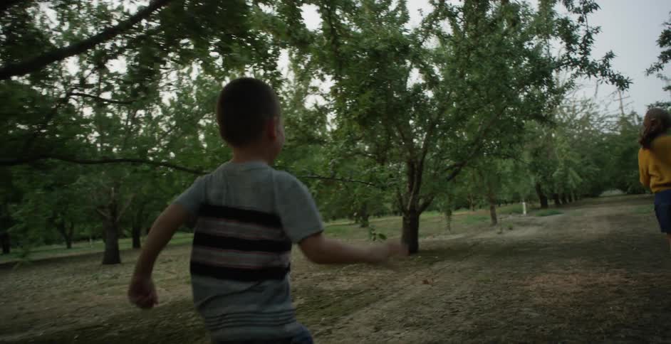 Boy wearing gray t shirt and jeans chases girl wearing yellow sweater and skirt in large garden at sunset