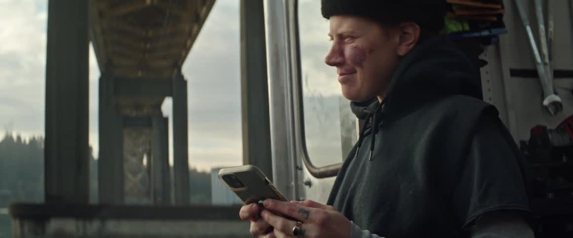 A woman is sitting outside in the doorway of a vehicle under a bridge with tools on the wall inside as she texts on her phone and smiles