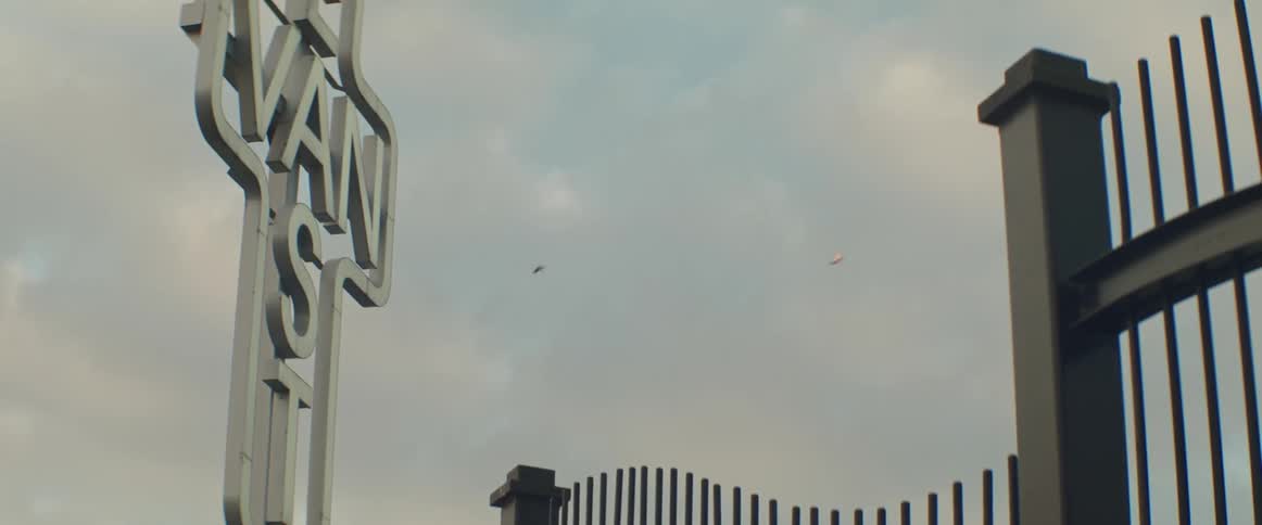 A fence and white East Van sign against a cloudy sky with birds flying across
