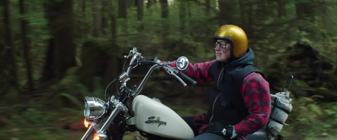 A woman in a gold helmet riding a motorcycle one handed through a forested area
