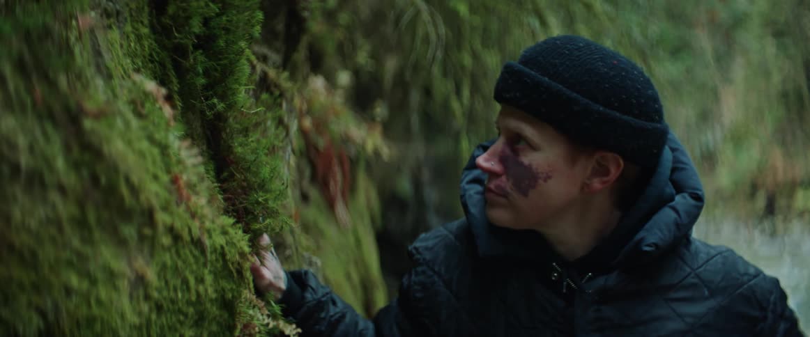 A woman wearing a coat and knit cap puts her hand on moss growing on a tree as she walks through the woods