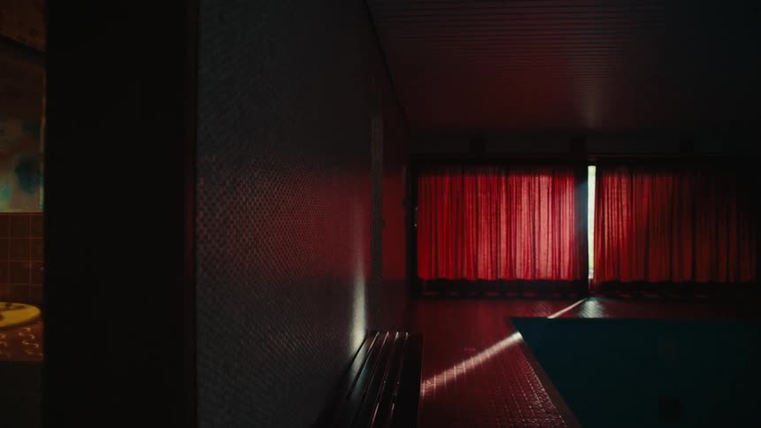 A green rug is on the hardwood floor as the sun shines through the red curtains and lights are on over the bathroom sink