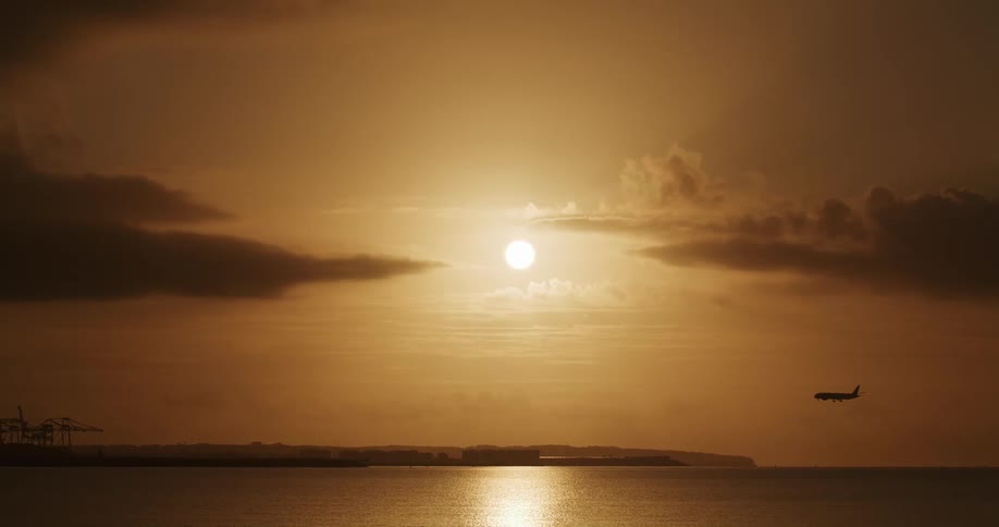 Sun shines brightly in red sky reflecting off the water as airliner approaches loading cranes
