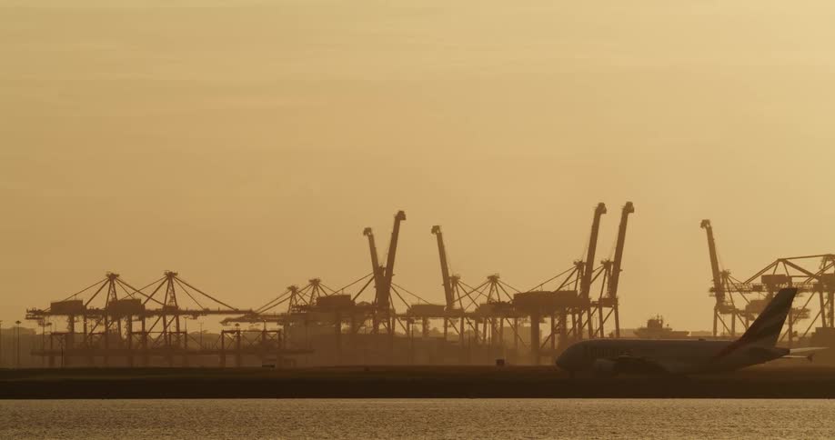 Airplane slowly moves along runway between cranes and water under a red sky