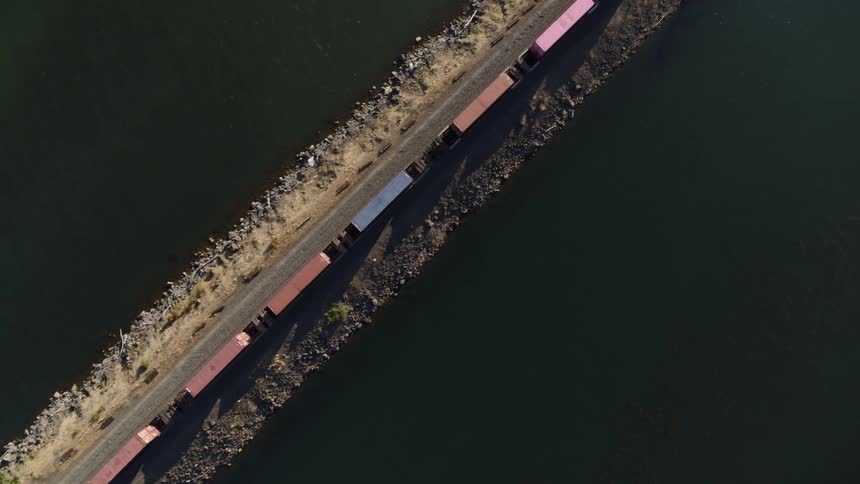 A freight train carrying shipping containers travels along a narrow causeway surrounded by water