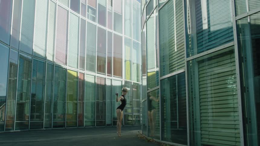 Female dancer dances between glass paneled buildings while viewing her reflection