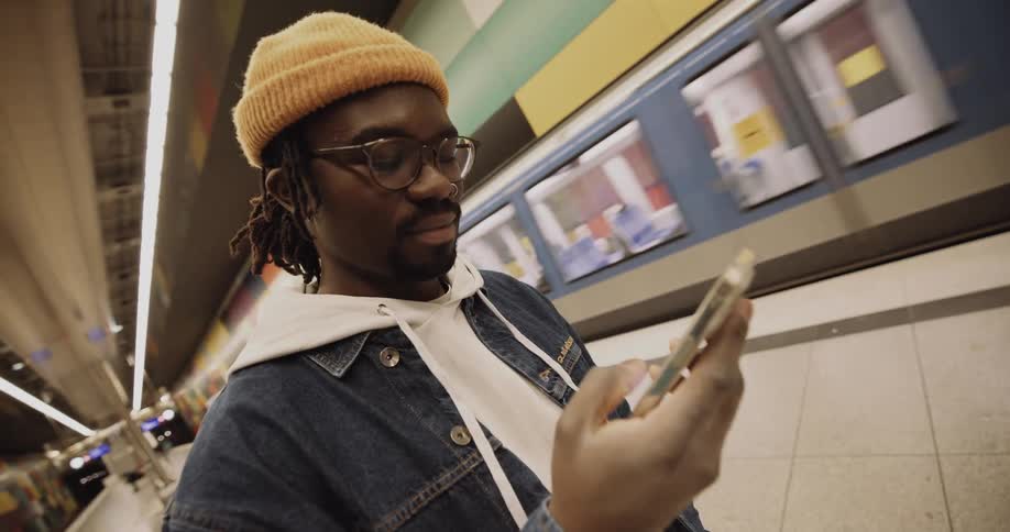 A man wearing a yellow hat standing next to a subway pulls out his phone and looks at it