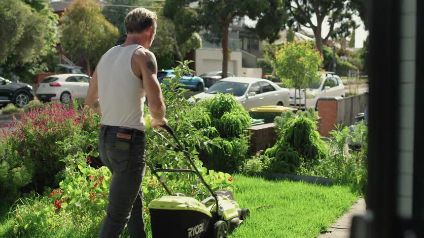 A man is outside mowing the lawn near the street and a flower bed