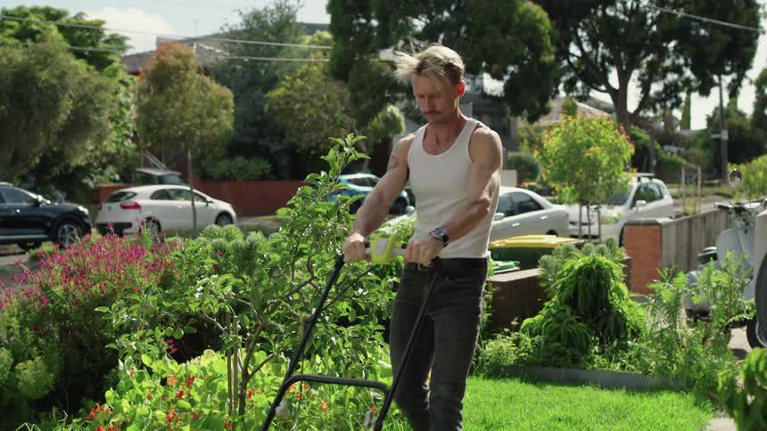 A man mowing a small plot of grass in a white tank top and black jeans