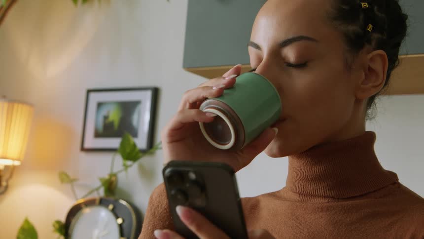 A woman wearing a turtleneck sweater drinking out of a small mug holding her cell phone