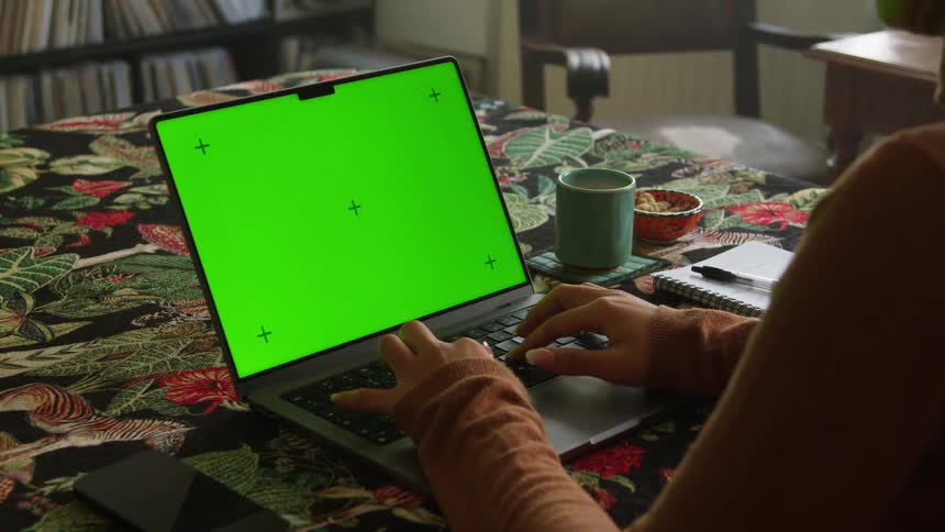 A woman is sitting at a table typing on a laptop computer with a green screen