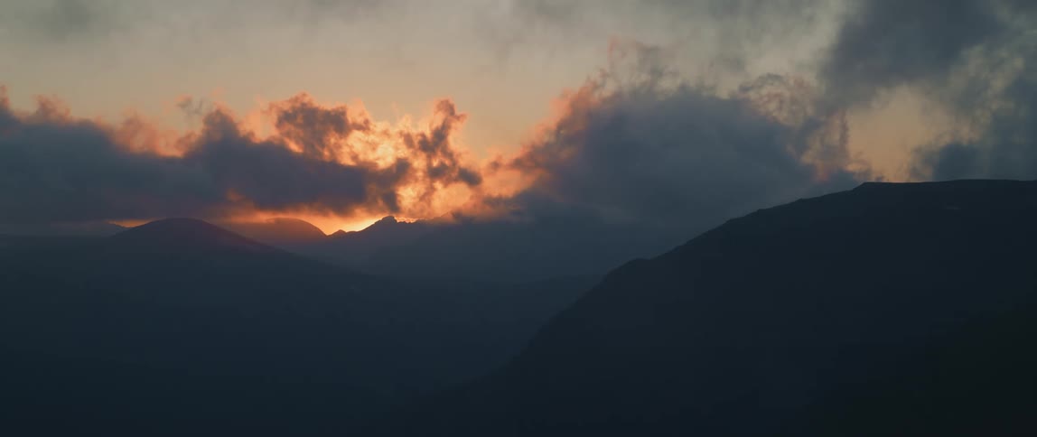 The sun rises through dense clouds over a mountain