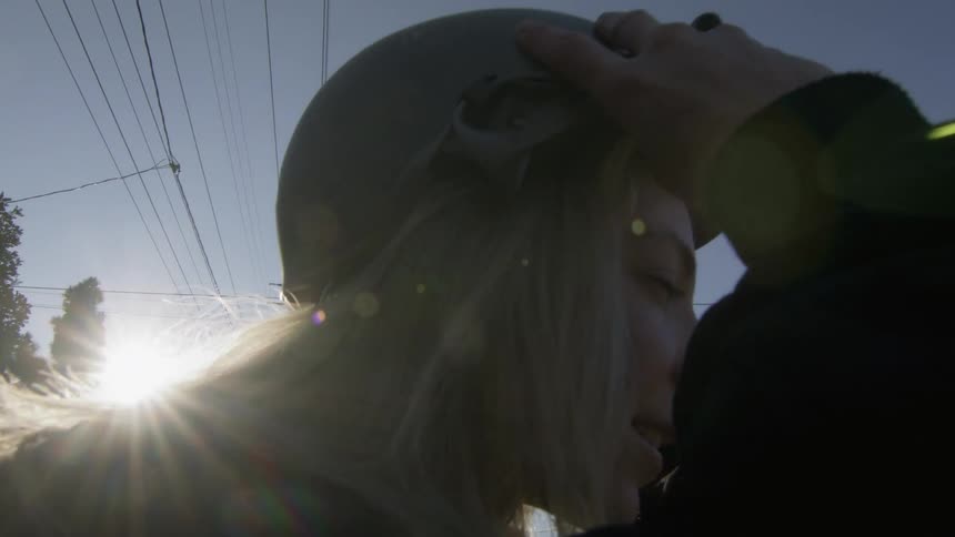 Sun shines behind a young blonde woman putting on a scooter helmet at morning