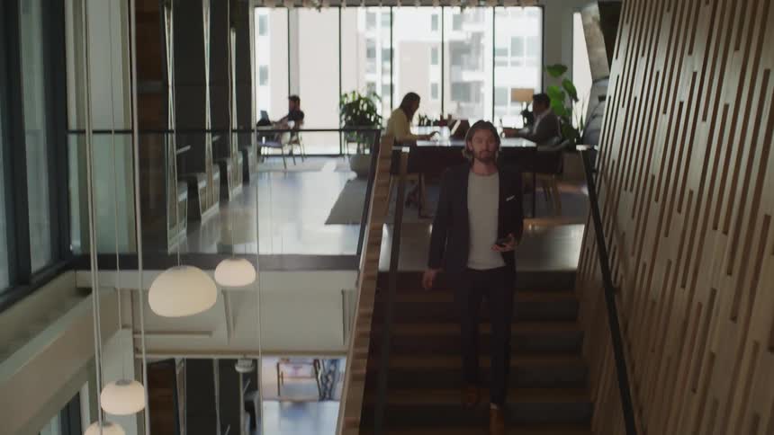 Man checks his phone while walking down stairs while coworkers sit at their desks working