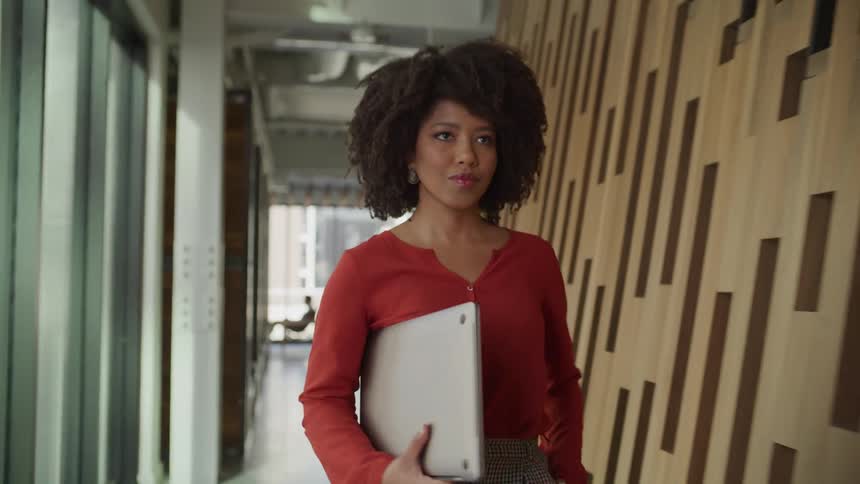 A woman wearing a red shirt holding a laptop walks briskly along a wooded wall