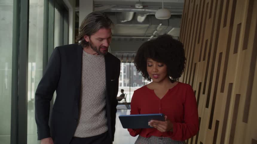 A woman carrying a tablet speaks with a man as they walk together through a corridor in an office building