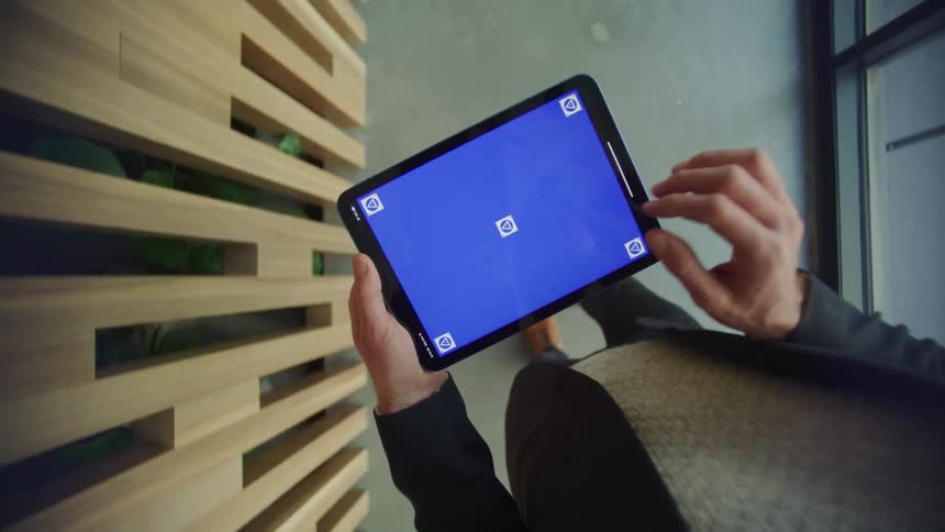 Man wearing a blazer jacket and brown sweater uses tablet while walking in office building