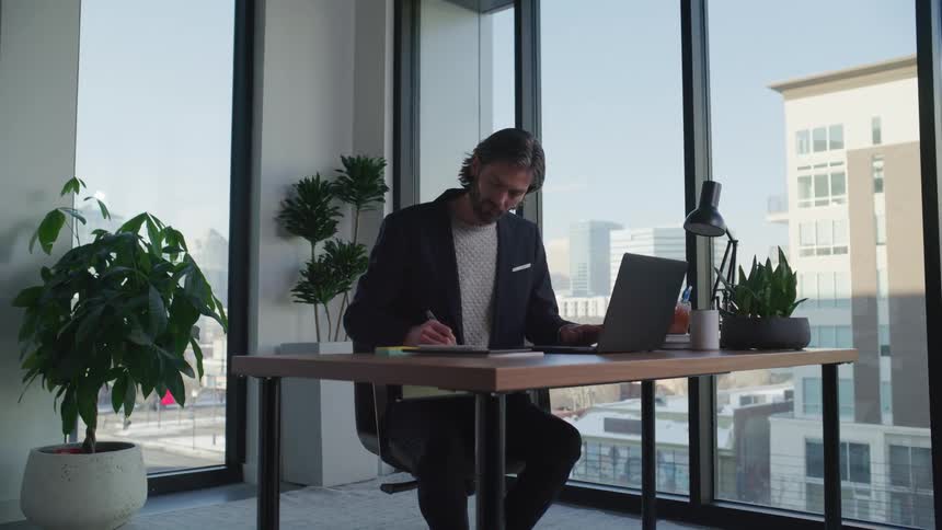 A man sitting at his desk in front of a laptop while he writes something down