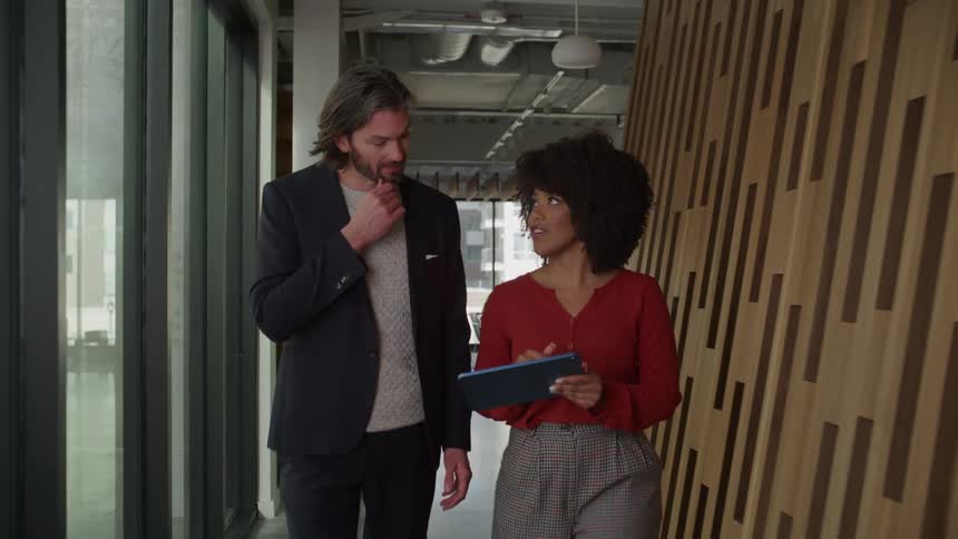 Businessman wearing blazer jacket and sweater walks next to businesswoman in red blouse and gray pants using tablet in office building