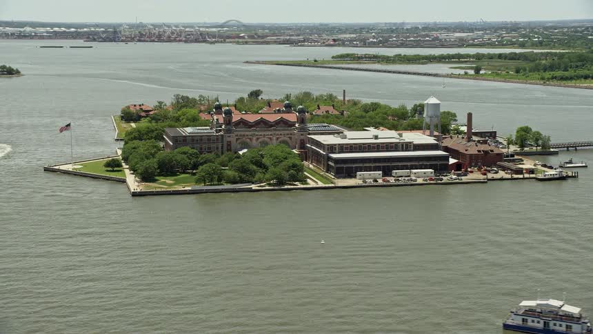 Aerial footage of Ellis Island in New York Harbor during a sunny day showcasing the historic landmark and surrounding buildings