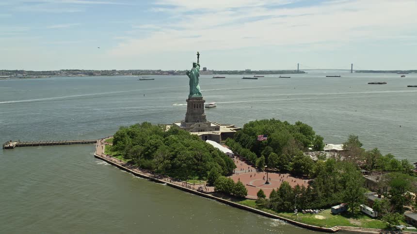 Aerial footage of Statue of Liberty orbiting around New York Harbor with people walking near the historic landmark under partly cloudy skies