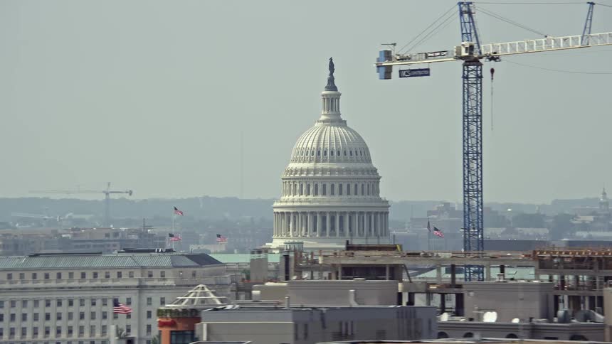 An aerial view of the iconic United States Capitol dome visible above office building rooftops in hazy Washington DC during summer