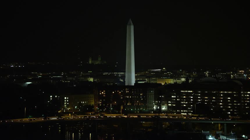 Approaching the Washington Monument and city lights an aerial view captures the historic landmarks and office buildings of Washington DC at night