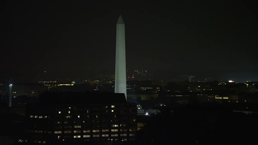 A mesmerizing aerial view of the Washington Monument draped in American flags at night surrounded by the clear historic landmarks of Washington DC