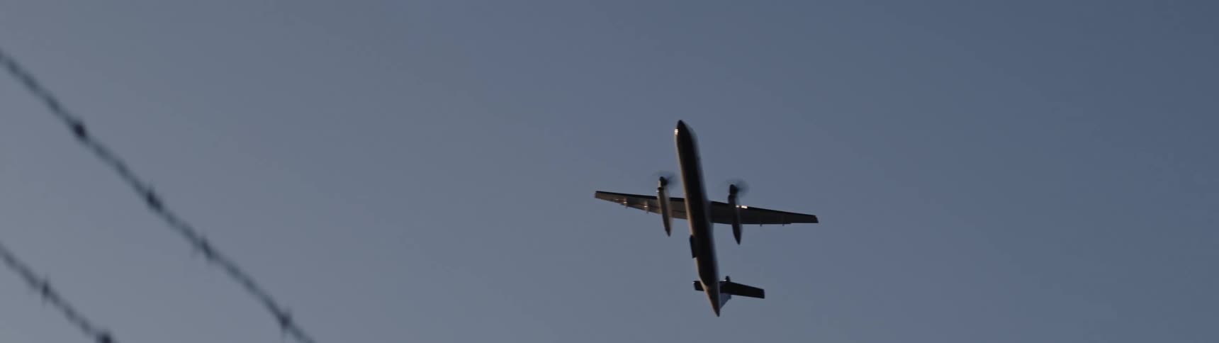 An airplane flies high above barbed wire on top of a chain linked fence as it comes in for a landing