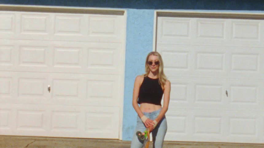 A blonde woman wearing a black cut off stands holding a skateboard in front of garage doors