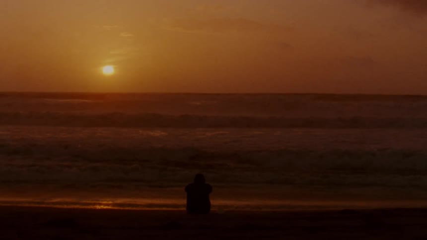 Silhouette of a person sitting on the beach looking at the sunset in a deep orange sky as the waves roll into shore