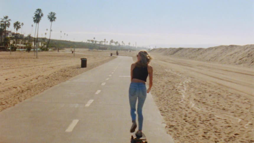 Sun shines over a young fit woman riding a skateboard on a road on a beach at daytime