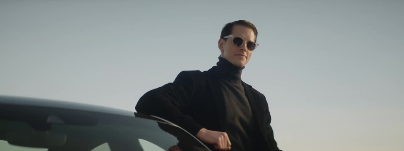 A man wearing sunglasses poses for a portrait with his elbow on the roof of a car