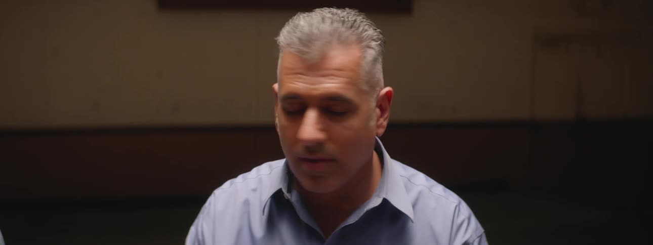 A man talking to two people offscreen before looking towards the camera in surprise