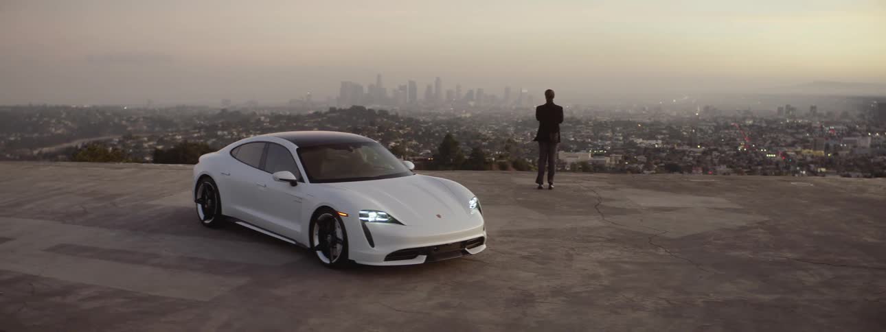 A man wearing a black blazer and sunglasses gets out of a white sports car parked overlooking neighborhoods and he walks to the edge looking at the big city skyline in the distance as he answers his phone