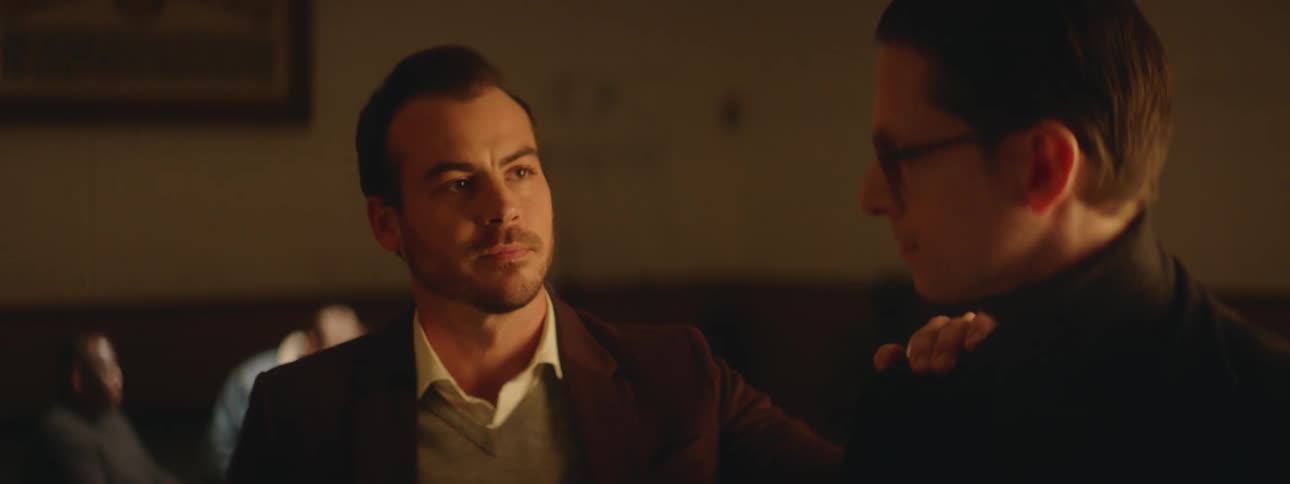A small group of men are sitting in a room and one man is standing as another man walks over to him and puts his hand on his shoulder as he takes something from inside his jacket and hands it to him