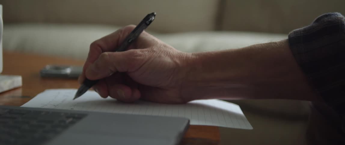 A persons hand writing with a pen on lined paper next to a laptop on the table