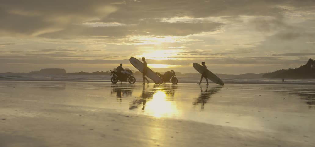 Surfers carry surfboards on a beach at sunset as they walk by two parked motorcycles