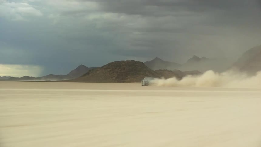 A black sports car leaves a trail of dust as it spins out in the desert under dark storm clouds and there are mountains in the distance