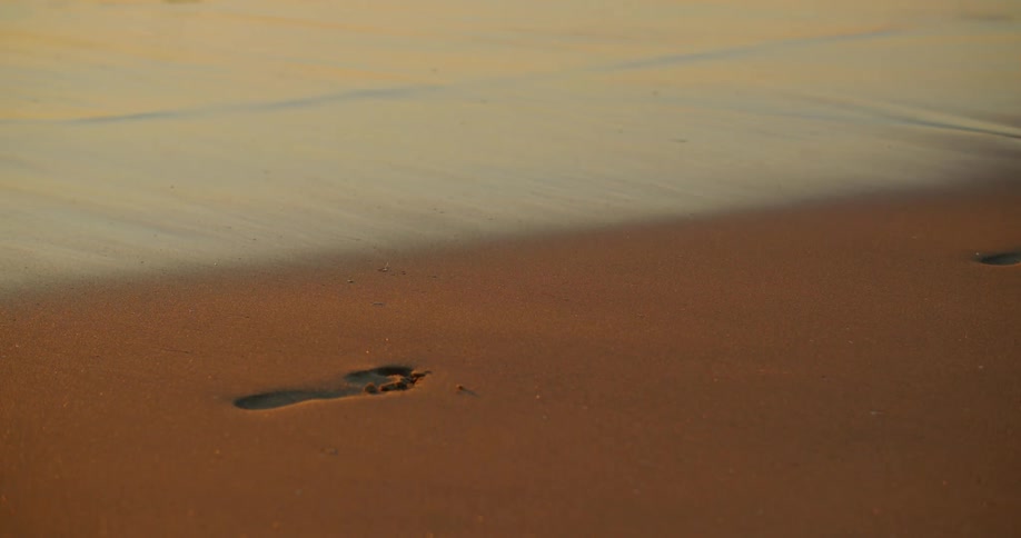 The sun shines upon sand on a beach as someone runs by barefoot and leave their footprints in the sand