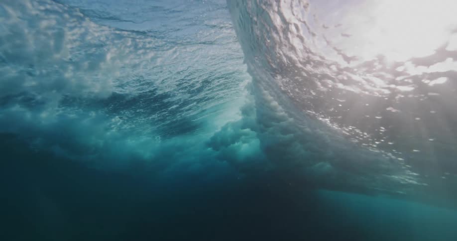 Rays of sunlight shine underwater and reflect on the ripples as a wave rolls overhead