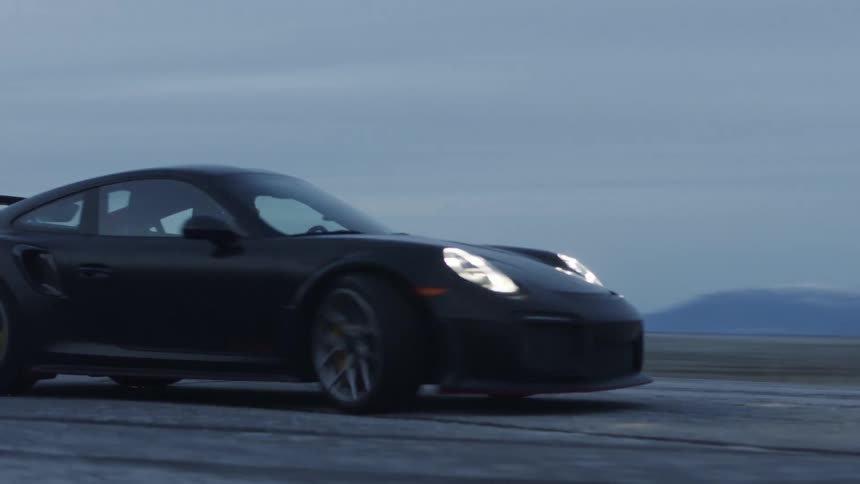 A dark sportscar travels fast and spins around on a road near mountains as the moon shines in the sky