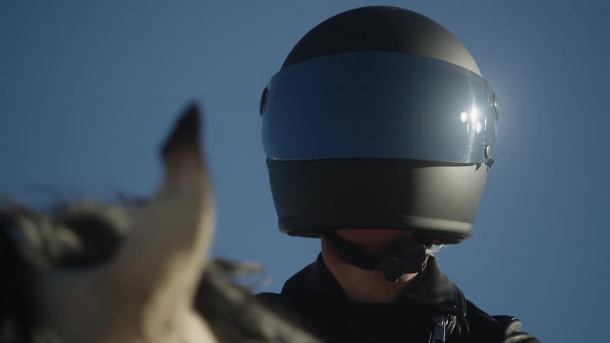 Woman in black helmet and leather jacket sits on white horse against the blue sky in the desert