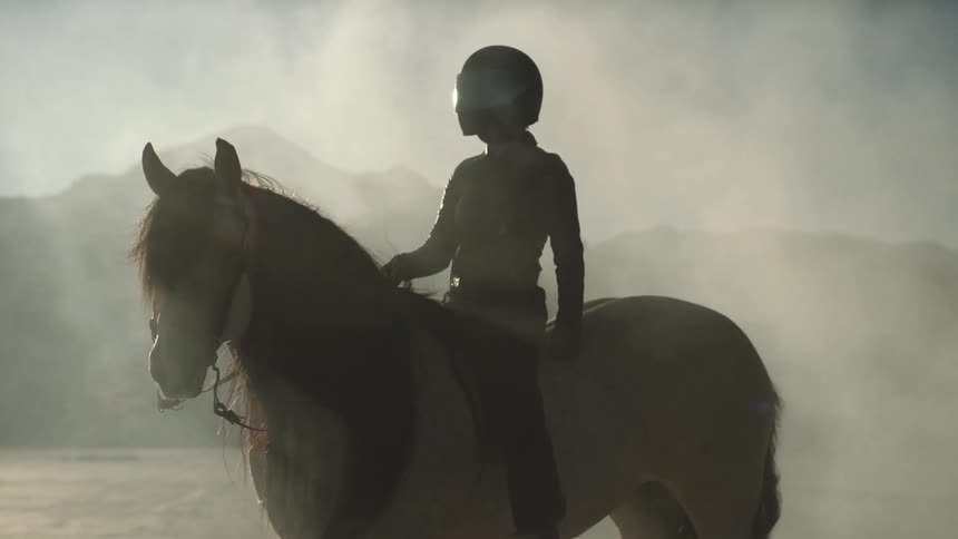 Person wearing motorcycle helmet sits on horse in dusty desert near mountains