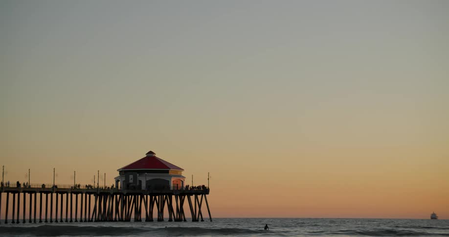 People move through water and walk along pier under a red sky with ship on the horizon