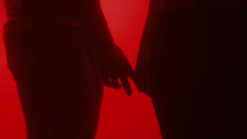 Man and woman circle around each other against a bright red background letting their hands touch