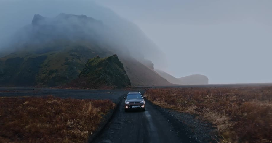 Brown car drives on dirt road near high rise mountain with fog