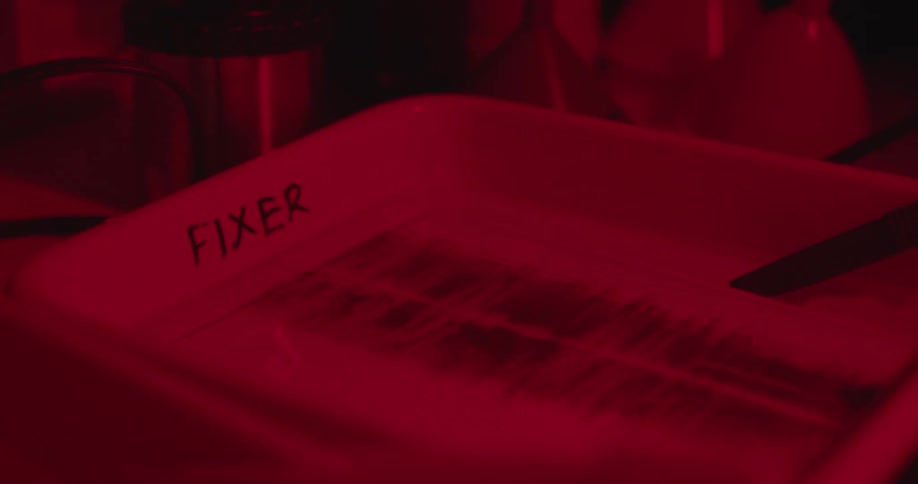 Red light glows in a darkroom as a photographer is developing film in a tray of liquid fixer